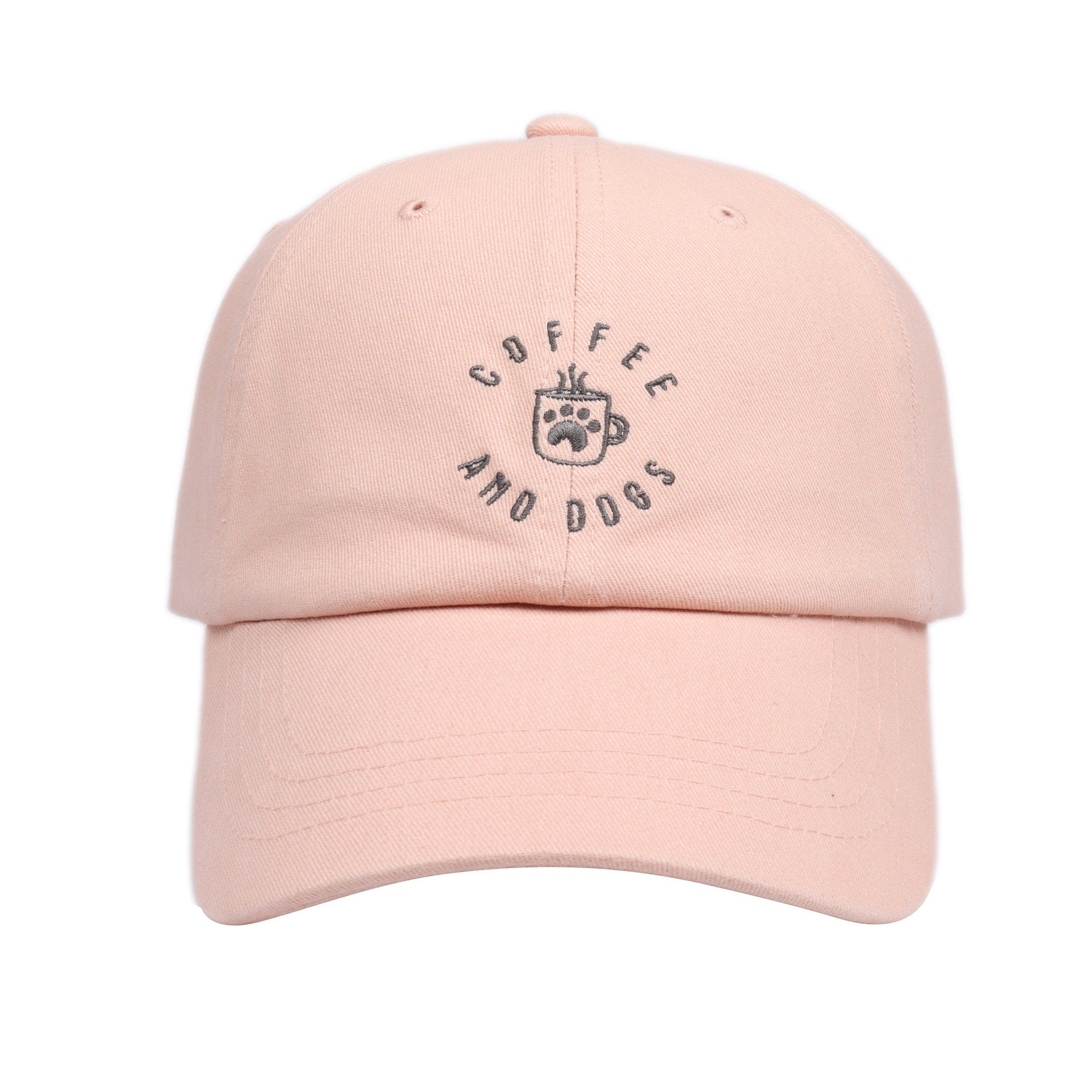 May include: A pink baseball cap with the text 'COFFEE AND DOGS' embroidered in gray on the front. The text is in a circle with a coffee cup and paw print in the center.
